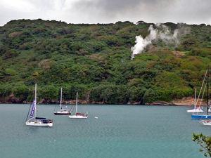 View of anchorage with six sailboats and hillside in background emitting steam